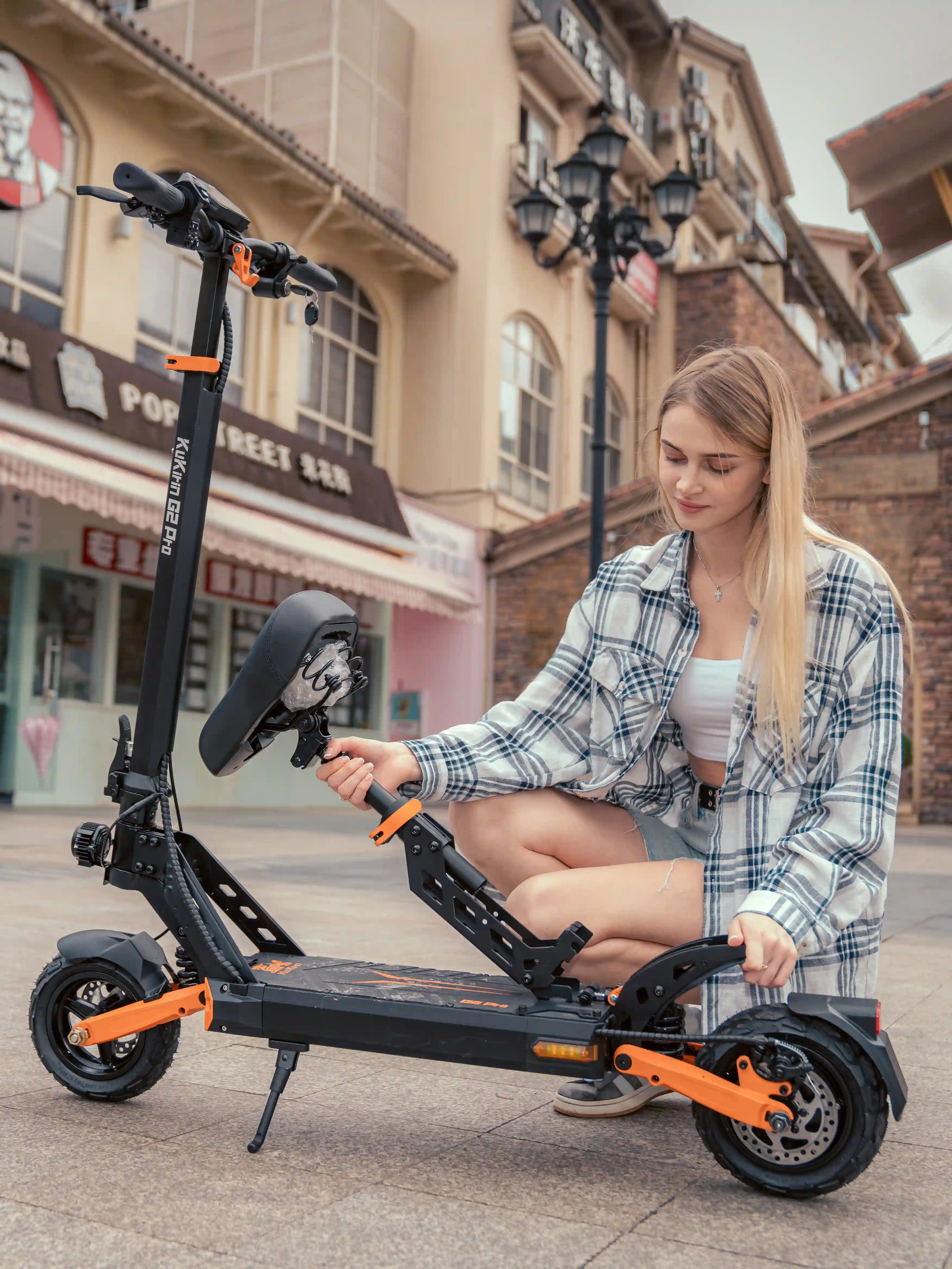 Lifestyle street photo of KuKirin G2 Pro foldable electric scooter, female user installing the detachable shock-absorbing seat, black and orange commuter e-scooter with dual spring suspension and 9-inch vacuum tires for city daily travel